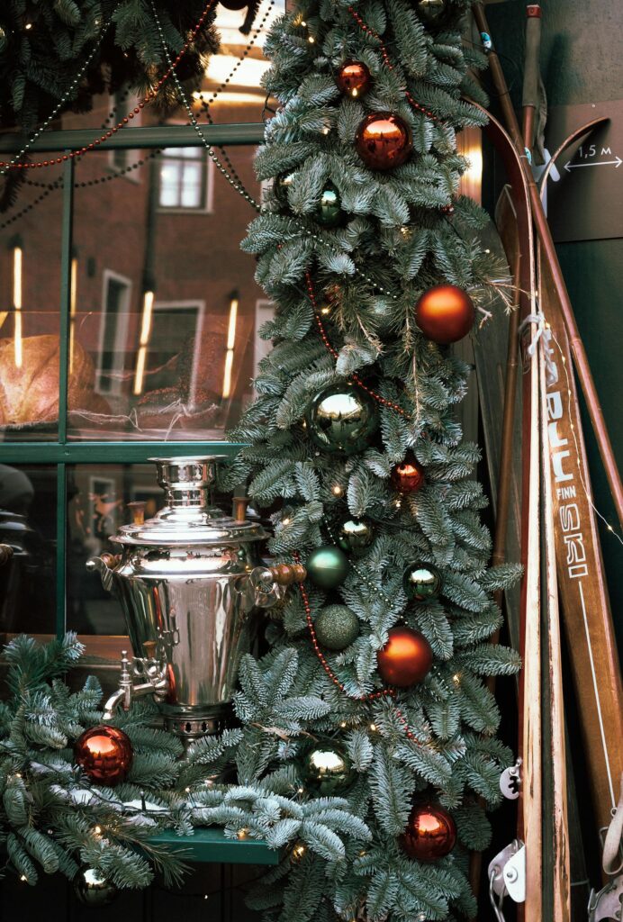 A charming Christmas window display adorned with colorful ornaments and a classic samovar amidst fir leaves.